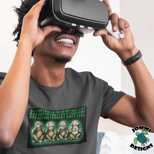 May include: A dark grey t-shirt featuring a graphic of four monkeys covering their eyes, ears, and mouth. The monkeys are surrounded by green binary code. The text "01110010 01010100 10100001 01110011 01110011 01010100 10100001 01111111" is printed above the monkeys.