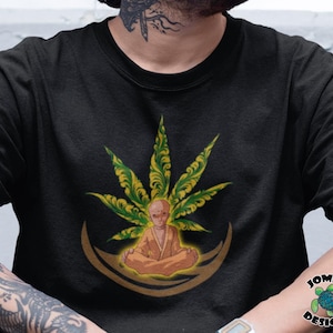 Puede incluir: Una camiseta negra con un diseño de hoja de marihuana verde y una figura meditando en el centro. La figura lleva una túnica amarilla y tiene una tez marrón claro. El diseño está rodeado por una forma de media luna dorada.