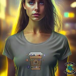 Puede incluir: Una camiseta gris con un gráfico de una taza de café con el texto "Ransom Demands 1 Cup of Coffee For 1 Hostage". El texto está en una fuente de estilo nota de rescate colorida. La camiseta la lleva puesta una persona con el pelo largo y castaño y ojos azules. El fondo está desenfocado y fuera de foco.