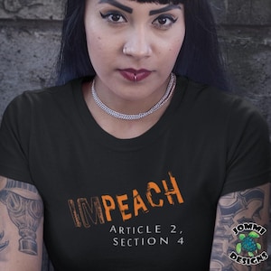 May include: Black t-shirt with the word "IMPEACH" in orange, with "ARTICLE 2, SECTION 4" below. The shirt is worn by a person with dark hair and a silver necklace.