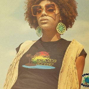 May include: A woman wearing a black t-shirt with a colorful graphic that says "Irie Vibe Chaser" and a brown suede vest. She is wearing sunglasses and green earrings.