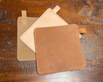 May include: Three leather pot holders in different shades of brown. The pot holders are stitched around the edges and have a loop for hanging.
