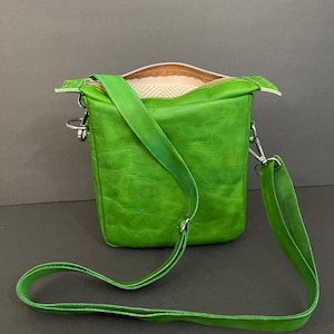 Puede incluir: Un bolso bandolera de cuero verde brillante con cierre de cremallera y una correa larga ajustable. El bolso tiene un forro de tela marrón claro.