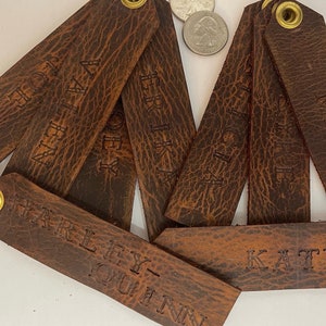 May include: A set of eight brown leather keychains with brass rings and personalized names, including Harley-Quinn, Kat, Chloe, Ava, Trix,  Africa, and  Sine.
