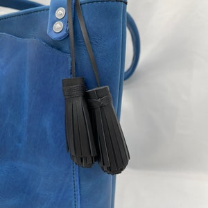 Può includere: Una borsa tote in pelle blu con cinturini e nappine in pelle nera. La borsa ha due bottoni a pressione in metallo argentato sul cinturino.