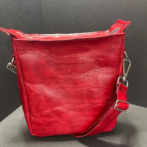 Puede incluir: Un bolso bandolera de cuero rojo con una correa larga ajustable. El bolso tiene una superficie texturizada y un cierre de cremallera.