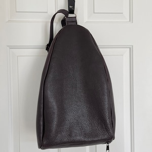 May include: A brown leather backpack with a zip closure. The backpack is hanging from a hook on a white door.