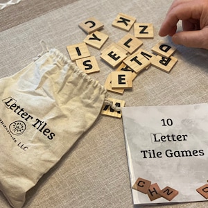 Può includere: Un sacchetto di tela beige con la scritta "Letter Tiles" e un logo ad albero, accompagnato da tessere di lettere in legno e un libretto intitolato "10 Letter Tile Games". Le tessere sono sparse su una superficie neutra, con una mano che le tocca.