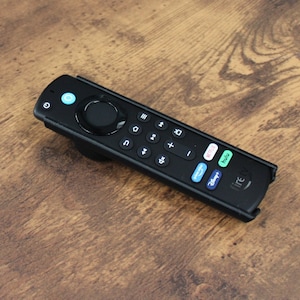 May include: Black remote control with various buttons, including a circular navigation pad and volume controls. The remote features a power button and app shortcut buttons. It is on a wooden surface.