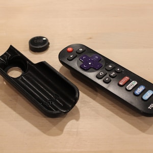 May include: Black 3D-printed remote control holder and a black remote control with purple directional buttons. A small black Tile tracker is also present. The remote is a TCL Roku TV model.