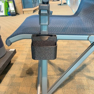 K&ouml;nnte beinhalten: Nahaufnahme eines schwarzen Getr&auml;nkehalters, der an einem blau-grauen Klappstuhl befestigt ist. Der Getr&auml;nkehalter besteht aus einem strukturierten, gesprenkelten Material. Der Stuhl hat eine blaue Netzbespannung. Das Wort "YETI" ist auf dem Rahmen zu sehen.
