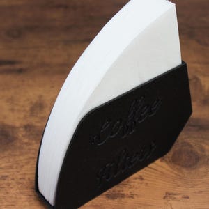 May include: A black coffee filter holder with the words "Coffee Filters" embossed on the front. The holder is filled with white paper coffee filters. The holder is on a wooden surface.