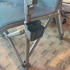 K&ouml;nnte beinhalten: Ein schwarzer Kunststoff-Getr&auml;nkehalter, der am Arm einer Klappstuhls befestigt ist. Der Stuhl hat eine blaue Netzbespannung und ein graues Metallgestell. Der Getr&auml;nkehalter ist strukturiert und hat eine rechteckige Form mit gew&ouml;lbter Oberseite.