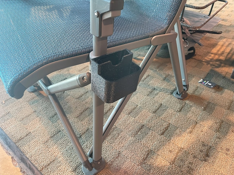 K&ouml;nnte beinhalten: Ein schwarzer Kunststoff-Getr&auml;nkehalter, der am Arm einer Klappstuhls befestigt ist. Der Stuhl hat eine blaue Netzbespannung und ein graues Metallgestell. Der Getr&auml;nkehalter ist strukturiert und hat eine rechteckige Form mit gew&ouml;lbter Oberseite.