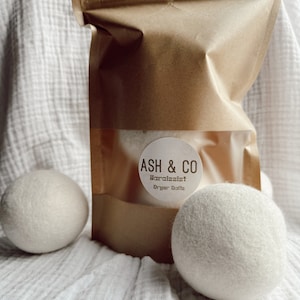 May include: A brown paper bag with a white label that says "ASH & CO Narcissist Dryer Balls". Two white dryer balls are in front of the bag on a white fabric surface.