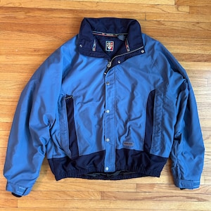 May include: A blue and navy jacket with a high collar and a zipper closure. The jacket has a logo on the chest and dark navy panels on the sides and bottom. The jacket is made of a lightweight material.