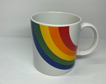 May include: White ceramic mug with a rainbow design. The rainbow arcs across the side of the mug in bands of blue, green, yellow, orange, and red. The mug has a white handle and a capacity of approximately 12 ounces.