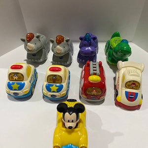 May include: A collection of VTech toys including a yellow car with Mickey Mouse, a red fire truck, a white police car, a white car with a star, a green dinosaur, a purple hippopotamus, and two gray rhinoceroses.