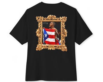 May include: A black t-shirt featuring a central graphic of a person holding a Puerto Rican flag, enclosed within an ornate gold-colored frame. The graphic includes a signature in the top left corner and text at the bottom of the frame.