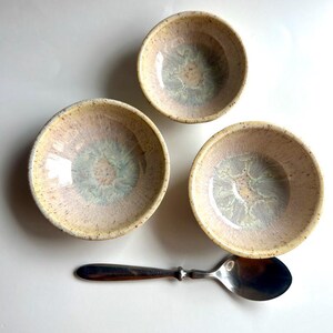 Op de afbeelding: Drie kleine keramische kommen met een gespikkeld, crèmekleurig exterieur en een lichtblauw en roze interieurontwerp. Een zilveren lepel ligt naast de kommen. De kommen hebben een rustieke, handgemaakte uitstraling.