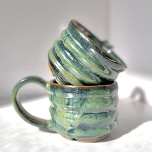 May include: Two stacked ceramic mugs featuring a glossy, variegated green and blue glaze with a distinct ribbed, horizontal texture. The bottom mug has a curved handle, while the top mug is tilted, showcasing the interior and the dark rim.