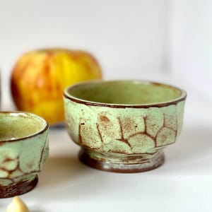 May include: Two small ceramic bowls with a light mint green glaze and a dark brown, textured, honeycomb-like pattern on the exterior. The bowls feature a wide, circular opening, a rounded body, and a solid, dark brown base that elevates the vessel.