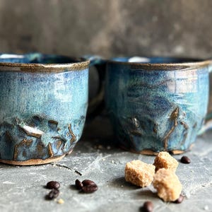 May include: Two handcrafted ceramic mugs with a blue and turquoise glaze. The mugs feature textured, sculpted details and brown rims. Coffee beans and sugar cubes are scattered around the mugs, suggesting a coffee theme.