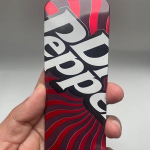 Puede incluir: Una etiqueta rectangular con un diseño rojo y negro con las palabras "Dr Pepper" en blanco, con una cuerda roja en la parte superior. La etiqueta tiene una estética retro y vintage.