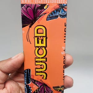 Può includere: Un pezzo di metallo piatto e rettangolare di colore arancione con la parola JUICED in grassetto giallo. La superficie è decorata con illustrazioni di farfalle colorate nei toni del viola, blu e giallo. Il testo include Vitamina B12 e dettagli.