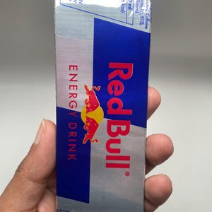 Può includere: Un'etichetta di lattina di bevanda energetica Red Bull, con sfondo argentato, una striscia blu e il logo Red Bull in rosso. L'etichetta include informazioni nutrizionali e le parole "ENERGY DRINK".