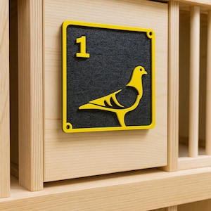 May include: A wooden pigeon coop with a yellow and black sign. The sign features a yellow outline, the number 1, and a yellow pigeon silhouette. The coop is made of light-coloured wood.