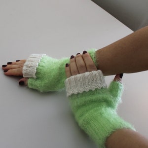 May include: A pair of green fingerless gloves with white knit cuffs. The gloves are made of a fuzzy yarn.