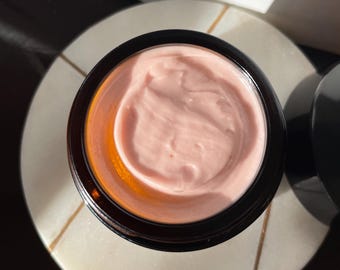 May include: A close-up of a glass jar with a pink cream inside. The jar has a dark brown lid and is sitting on a white surface.
