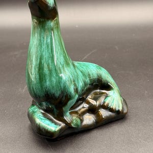 May include: A ceramic figurine of a seal in a seated position, featuring a glossy glaze with blended shades of deep emerald green and dark brown. The seal is perched on a textured, dark brown base that mimics a rocky surface with visible contours.