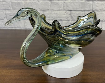 May include: A glass swan-shaped bowl with a swirl pattern in shades of green, blue, and brown. The bowl is sitting on a white pedestal.