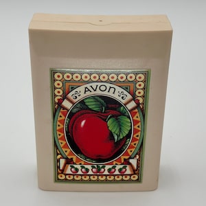 May include: A beige box with a red apple and green leaves on the front. The box has a gold border and the word "AVON" in a gold script font. There are small red apples around the bottom of the box.