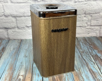 Puede incluir: Un bote de metal marrón y plateado con un patrón de veta de madera y una tapa plateada. El bote tiene la palabra "Swig-O-Mat" impresa en la parte delantera.