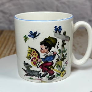 May include: A white ceramic mug with a blue rim features a colourful illustration of a child riding a rocking horse, a dog, and birds. The mug has a handle.