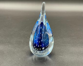 May include: A clear glass teardrop-shaped art piece with a vibrant blue interior. The blue section is filled with small, white air bubbles, creating a unique visual texture. The top of the piece tapers to a point.
