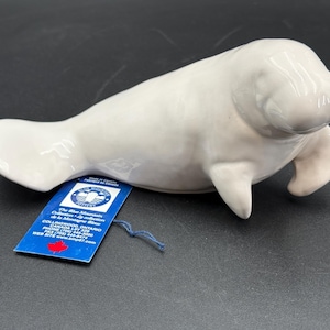 May include: A glossy white ceramic manatee figurine with a smooth, rounded body and flippers. Attached to the base is a blue rectangular tag with white text that reads Blue Mountain Pottery, Collingwood, Ontario, Canada, and a red maple leaf.