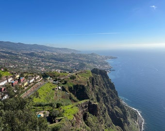 May include: A panoramic view from a clifftop overlooking a coastal town and the ocean. The town is nestled in the valley below, with green hills and a winding road leading up to the cliff. The ocean is a deep blue, with a few boats visible in the distance.