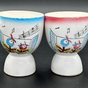 Può includere: Due porta uova in ceramica bianca con bordo blu e rosso. Ogni porta uova ha un design di note musicali con un uccello blu e rosso che suona strumenti.