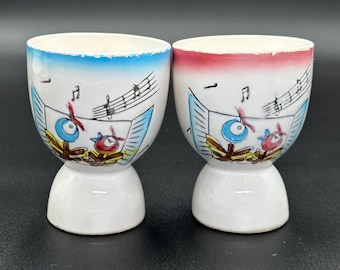 May include: Two white ceramic egg cups with a blue and red rim. Each cup has a musical note design with a blue and red bird playing instruments.