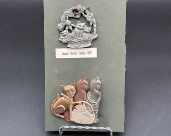 May include: Two vintage pewter cat pins. The top pin features a cat and kittens in a floral basket. The bottom pin shows three cats, one with a basket of kittens. The text "Seagull Pewter Canada 1987" is visible.