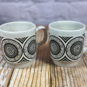 May include: Two white ceramic mugs with black and white geometric designs. The mugs have a rounded shape and a handle on the right side.