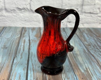 May include: A small, red and black ceramic pitcher with a handle. The pitcher has a glaze that looks like it has been dripped with black paint. The pitcher has a gold inscription that reads "'s alles do".