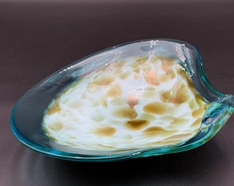 May include: A handcrafted glass bowl with a unique design. The bowl features a clear teal rim and a white interior with brown and gold speckles. The bowl has an organic, flowing shape, perfect for decorative use.