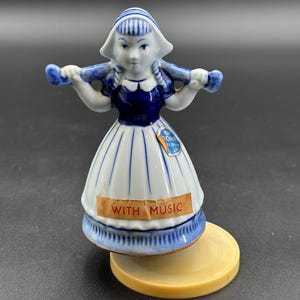 May include: A blue and white porcelain figurine of a Dutch girl wearing a blue and white dress and a blue and white hat. The figurine is holding a pair of wooden shoes and is playing music. The figurine is on a round base and has a label that says "With Music".