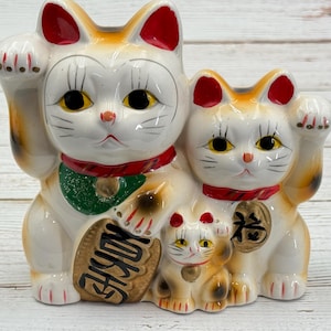 Może przedstawiać: Ceramiczna figurka rodziny Maneki Neko. Koty są białe z pomarańczowymi i czerwonymi akcentami, czarnymi oczami i podniesionymi łapami. Największy kot trzyma złotą monetę i transparent z japońskimi znakami. Koty noszą czerwone i zielone obroże.
