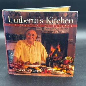 Peut inclure: Un livre de cuisine à couverture rigide intitulé Umberto's Kitchen: The Flavours of Tuscany par Umberto Menghi. La couverture présente une photographie d'une personne en chemise jaune dans une cuisine avec une cheminée, entourée de plats.